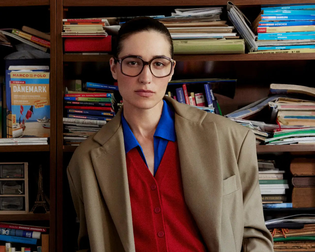 Model wearing large eyeglasses and brown coat standing in front of cluttered bookcase. Image credit: Andy Wolf