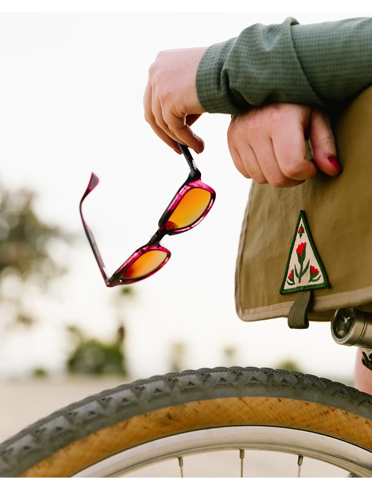 Person holding sunglasses next to a bicycle with a green jacket and floral patch. Image credit: Article One