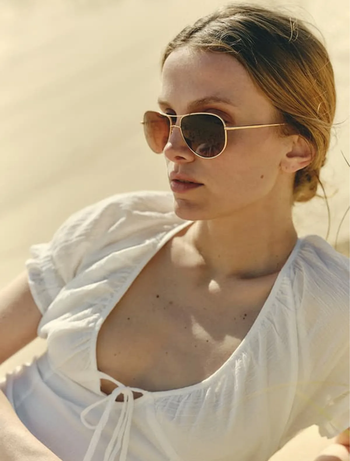 A woman in a white tie-front top wearing SALT. gold-framed aviator sunglasses with brown lenses, photographed in warm natural light against a soft beige background. Image credit: SALT