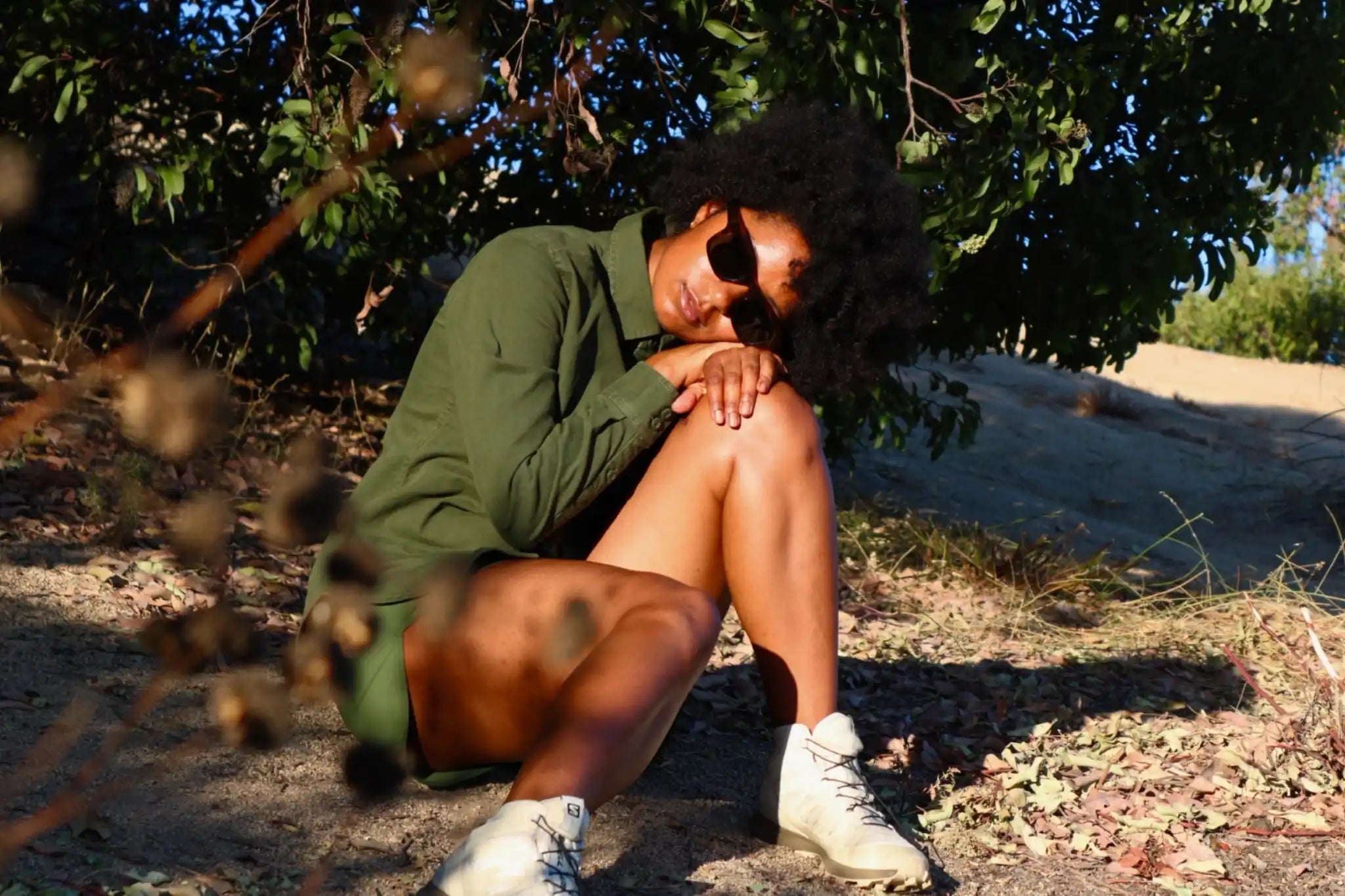 Model wearing Article One sunglasses sitting in the shade wearing a green jacket and white boots