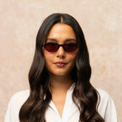 A woman with long, wavy dark brown hair wearing dark brown cat-eye sunglasses and a white collared shirt, set against a textured beige background.