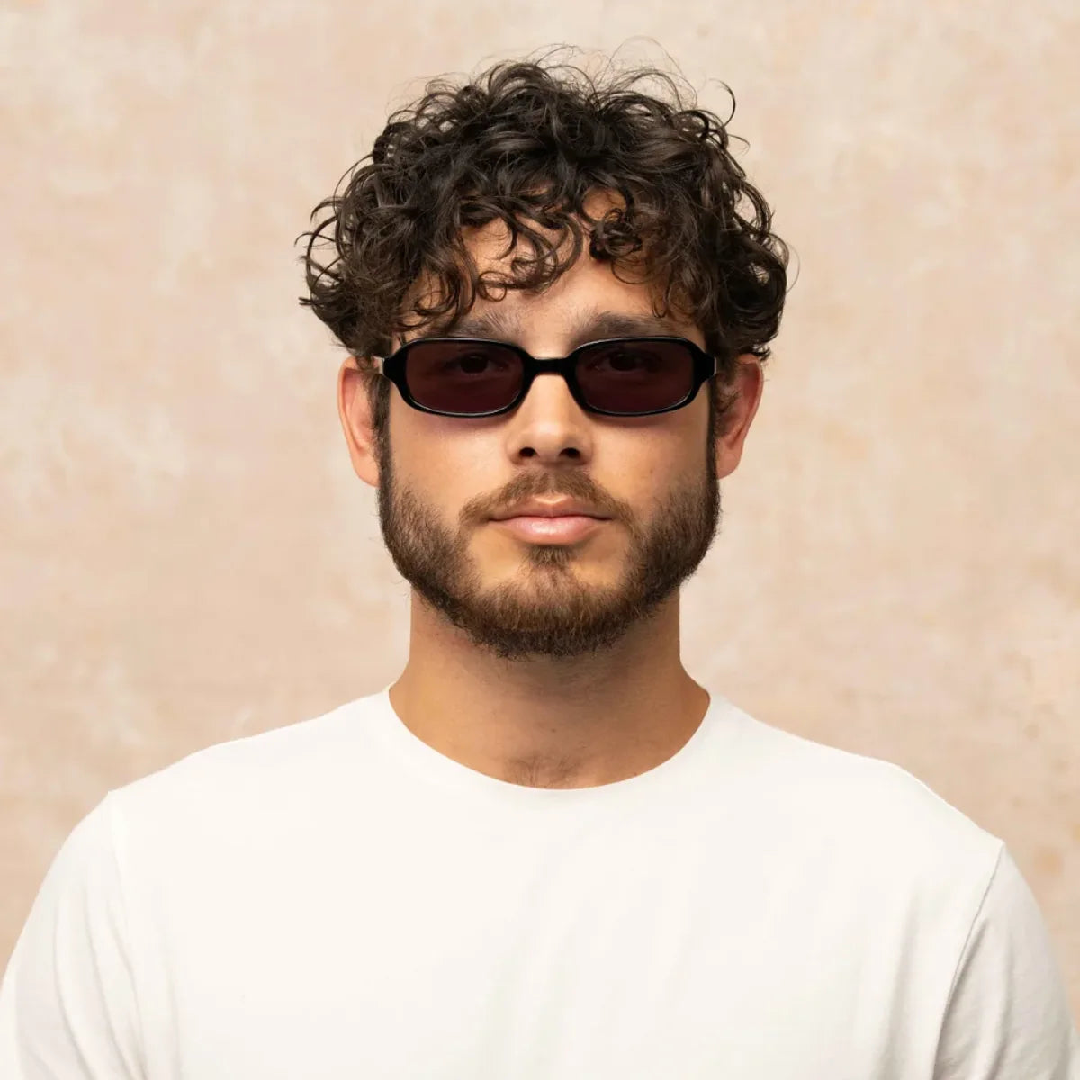 A man with curly dark hair and a beard wearing black oval Garrett Leight sunglasses and a white t-shirt, set against a textured beige background.