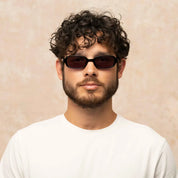 A man with curly dark hair and a beard wearing black oval Garrett Leight sunglasses and a white t-shirt, set against a textured beige background.